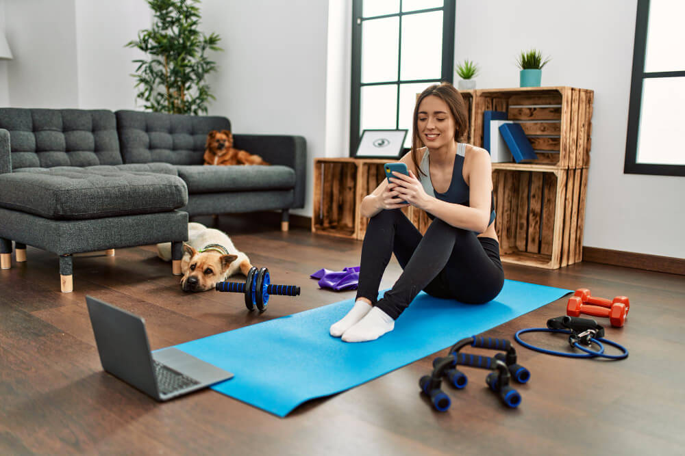 home-fitness - SaveTo Wishlist Young woman sitting on a blue yoga mat in a bright living room, smiling at her phone surrounded by workout equipment and two relaxed dogs.