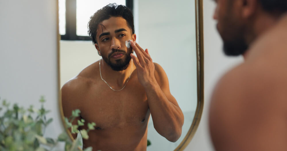 self-care - SaveTo Wishlist A shirtless man applies moisturizer to his cheek while studying his reflection in a bathroom mirror, natural light and plants nearby.