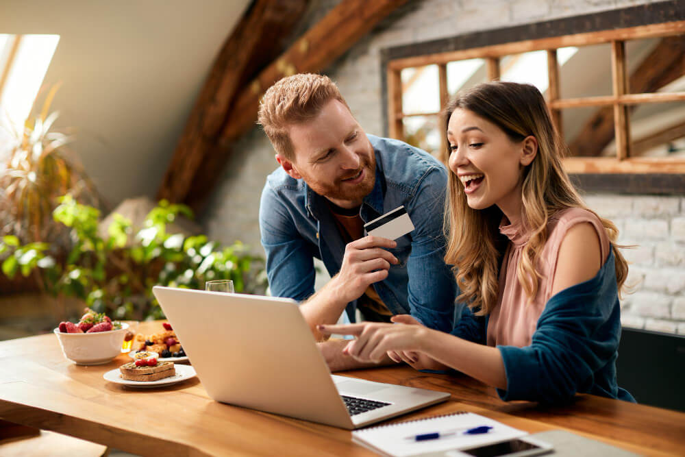 Smiling couple shopping online in a sunlit loft kitchen, man holding a credit card while woman excitedly points at the laptop screen.